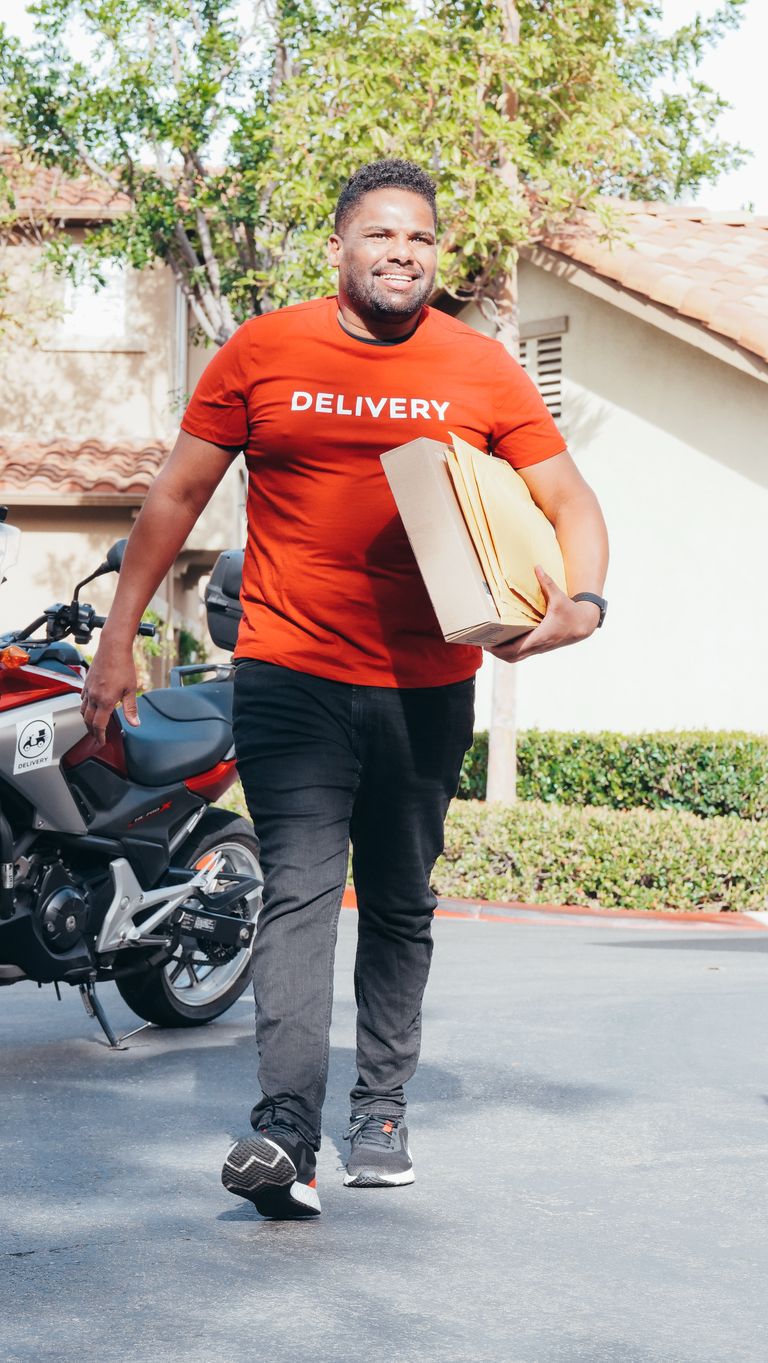 A cheerful delivery man carrying packages, walking outdoors in a sunny suburban area.